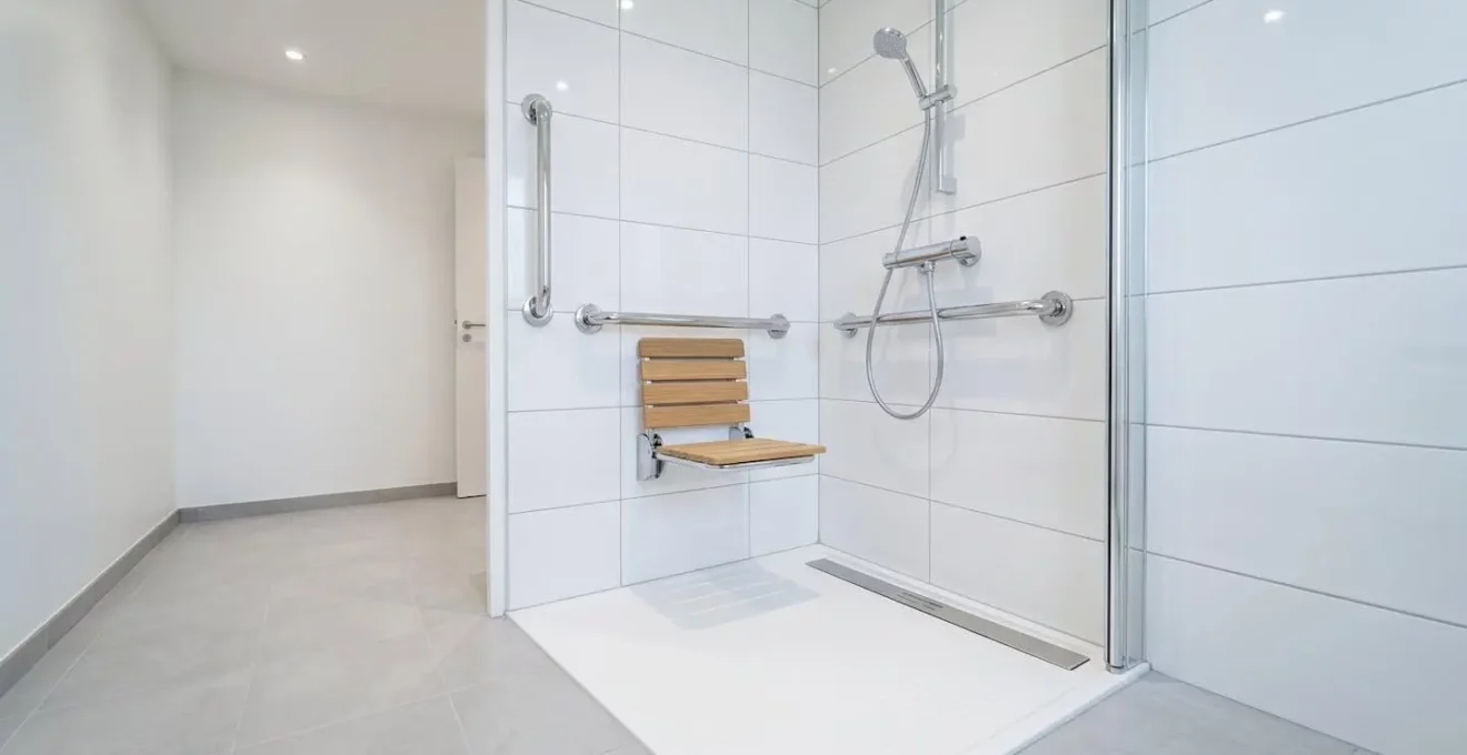 Douche à l'italienne plain-pied adaptée seniors avec barres d'appui chromées, siège rabattable en bois clair et carrelage antidérapant blanc et gris